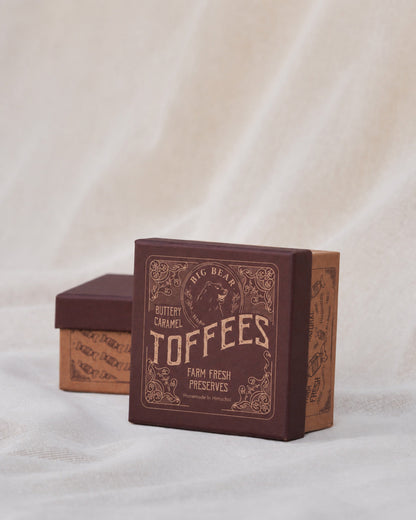 A square box of Big Bear Farms Caramel Toffees, shown in the foreground with its lid leaning against the side. A second box is visible in the background.