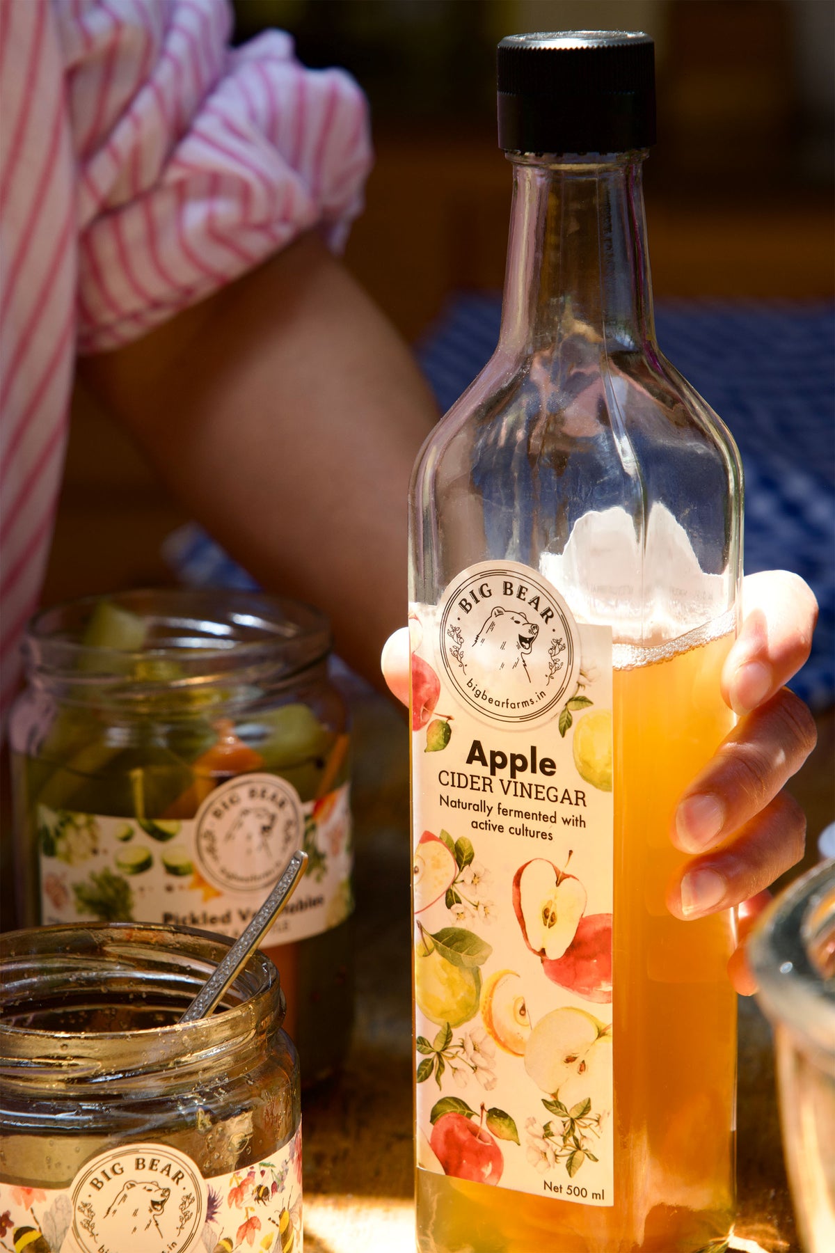 A hand holds a bottle of Big Bear Farms Apple Cider Vinegar in a warm, rustic setting. The clear glass bottle is filled with golden amber liquid and features a white label with illustrations of apples and citrus fruits.