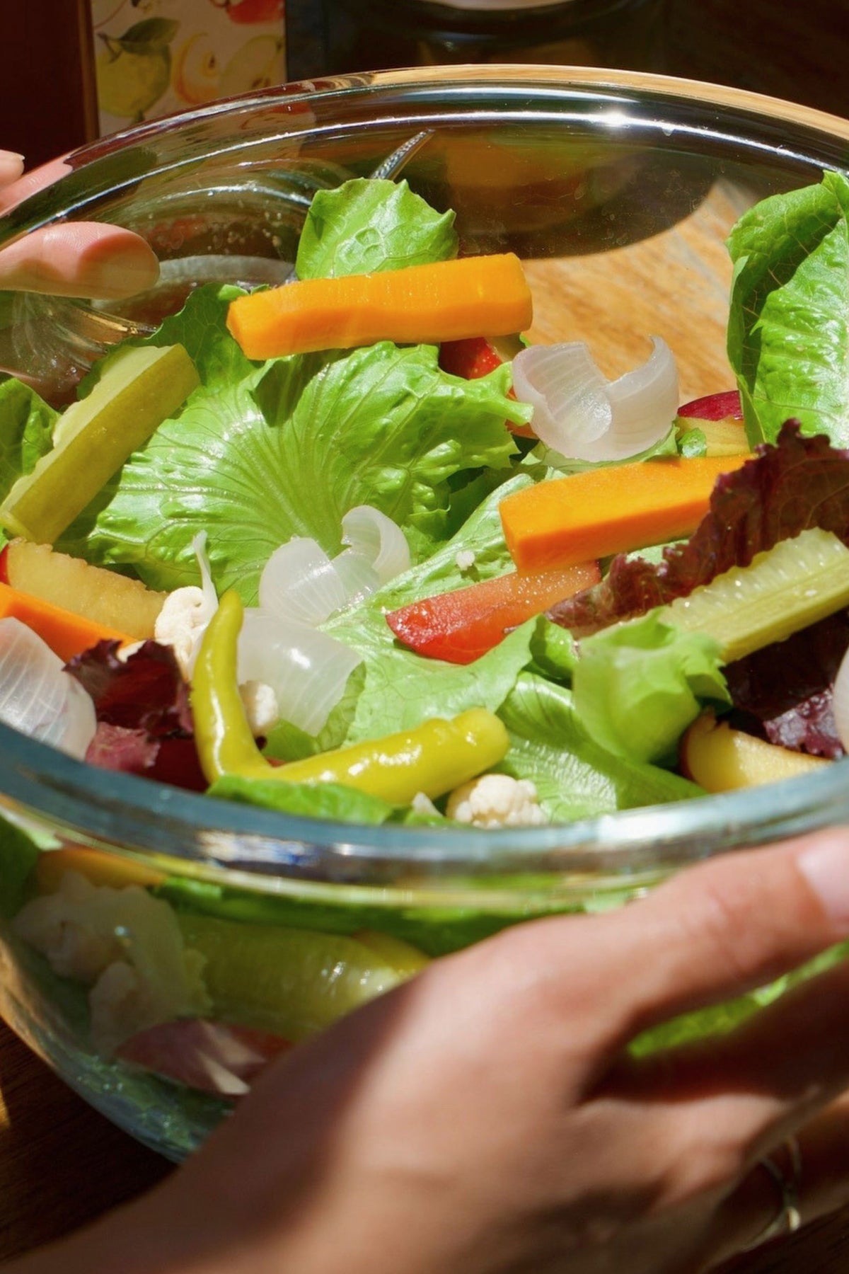 A close-up of a fresh mixed vegetable salad in a clear glass bowl, held by hands. The salad contains green lettuce, orange carrot sticks, red bell pepper pieces, yellow peppers, white onions and Big Bear Farms Apple Cider Vinegar dressing.