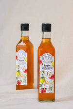 Two glass bottles of Big Bear Farms Apple Cider Vinegar with black caps, standing on a light beige fabric surface against a neutral beige background.