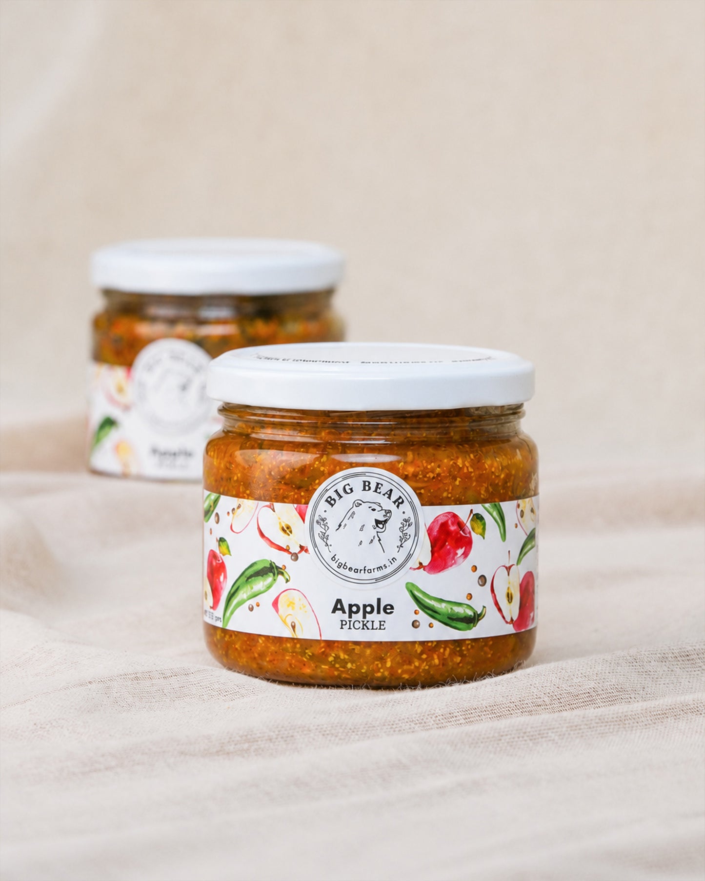Two glass jars of Big Bear Farms Apple Pickle arranged on a beige fabric surface, with the front jar in sharp focus and the back jar softly blurred.