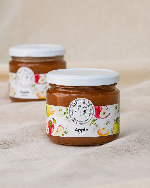Two glass jars of Big Bear Farms Apple Sauce on a beige fabric background, with the front jar in sharp focus and the rear jar softly blurred.