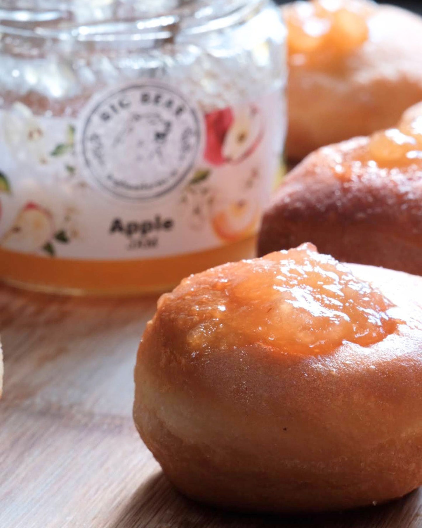 Close-up of golden-brown baked pastries topped with Big Bear Farms Apple Jam with a blurred jar of Big Bear Apple Jam in the background on a wooden surface.