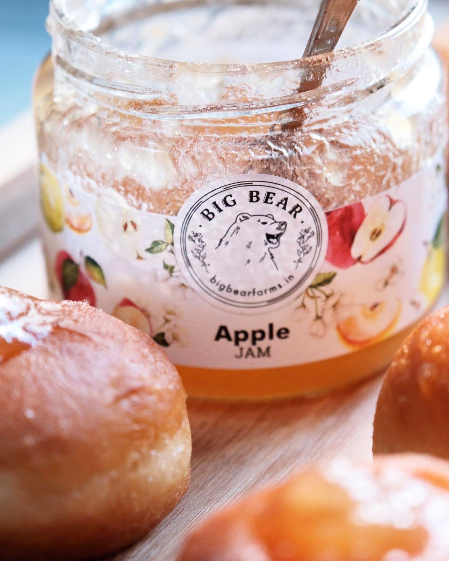 A glass jar of Big Bear Farms Apple Jam with a wooden spoon, surrounded by fresh apple pastries on a wooden surface.