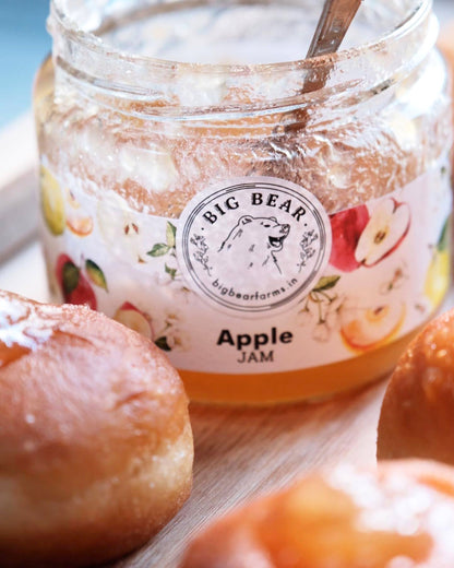 A glass jar of Big Bear Farms Apple Jam with a wooden spoon, surrounded by fresh apple pastries on a wooden surface.