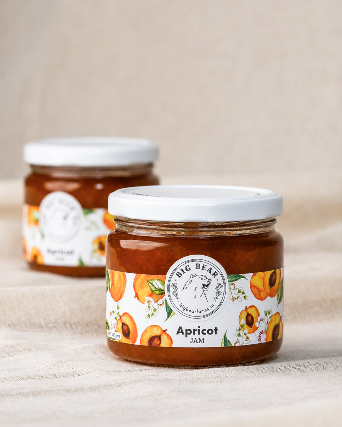 Two glass jars of Big Bear Farms Apricot Jam on a beige fabric background, with one jar in sharp focus in the foreground and the other blurred in the background.