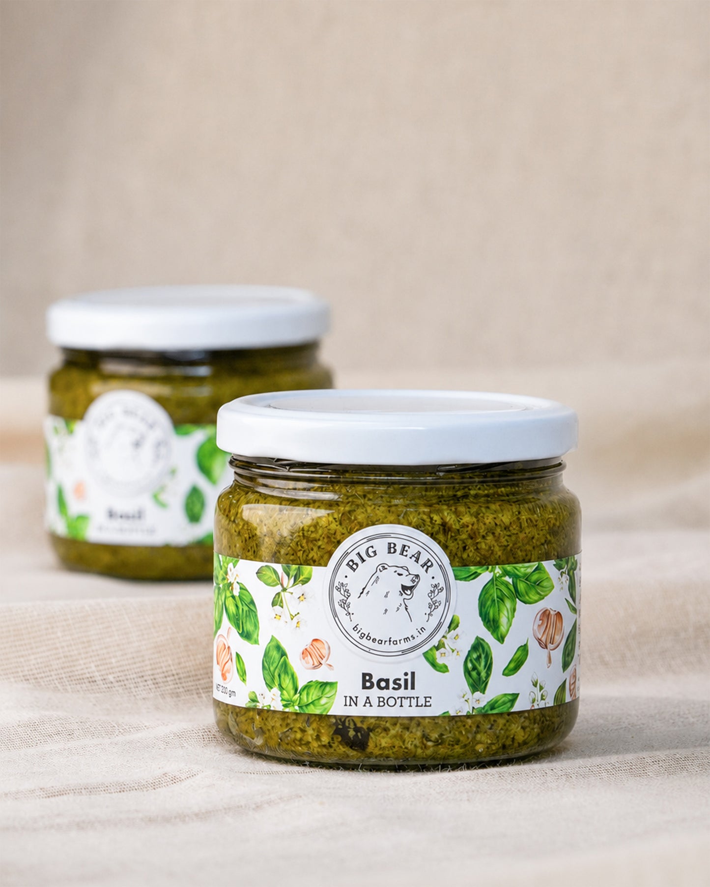 Two glass jars of Big Bear Farm Fresh Basil in a Bottle on a beige linen surface with a neutral beige background. The front jar is in sharp focus, showing the green pesto inside and a white label with green basil leaves, while the second jar is softly blurred in the background.