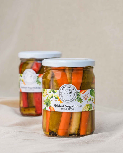 Two glass jars of Big Bear Farms Pickled Vegetables with white lids, set on a beige fabric surface against a neutral beige background.