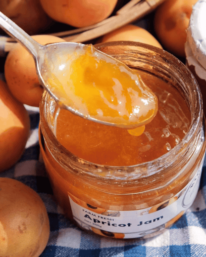 An open glass jar of Big Bear Farms apricot jam with a spoon lifting a glossy spoonful of the jam, surrounded by whole apricots and a blue-and-white checkered cloth.
