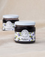 Two glass jars of Big Bear Farms Blueberry Preserve with white lids and blueberry fruit label, set on a beige fabric background.