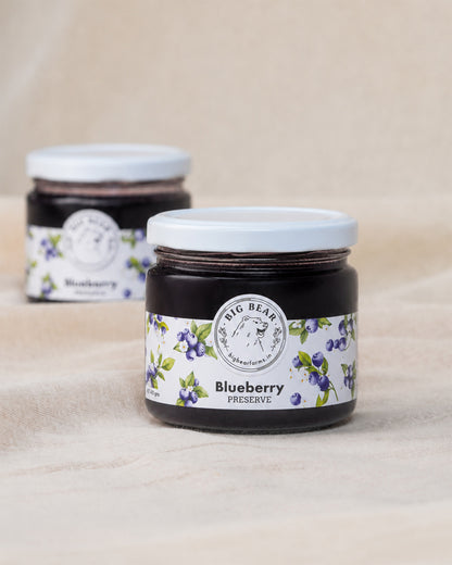 Two glass jars of Big Bear Farms Blueberry Preserve with white lids and blueberry fruit label, set on a beige fabric background.