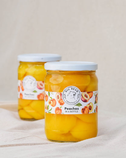 Two glass jars of Big Bear Farms Fresh Peaches in a Bottle, filled with yellow-orange peaches and set on a light beige fabric surface against a neutral beige background.