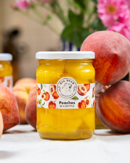 A jar of Big Bear Farms Fresh Peaches in a Bottle spread on a white surface, surrounded by fresh peaches and a blurred floral background.