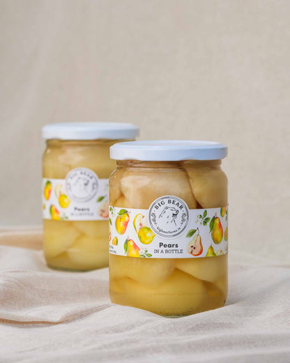 Two glass jars of Big Bear Farms Pears in a Bottle, filled with sliced pears and topped with white lids, set on a beige fabric surface against a neutral beige background.