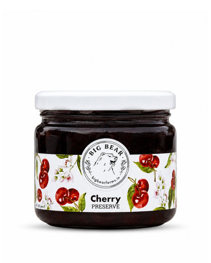 A glass jar of Big Bear Farms Cherry Preserve with a white lid and a white label decorated with red cherries and green leaves, set against a plain white background.