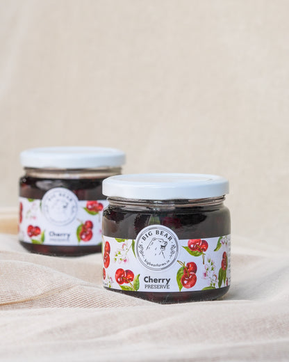 Two glass jars of Big Bear Farms Cherry Preserve with white lids and white labels featuring red cherries, set on a beige fabric surface.