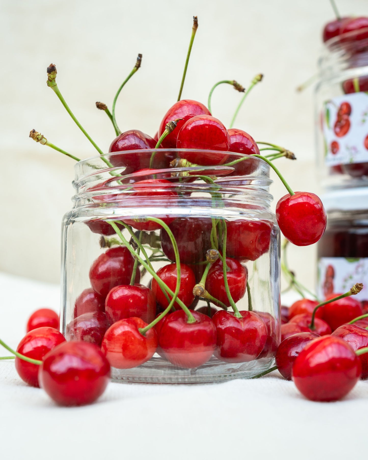 A clear Big Bear Farms glass jar filled with bright red cherries with green stems, set on a light surface with more cherries scattered around and a blurred jar in the background.