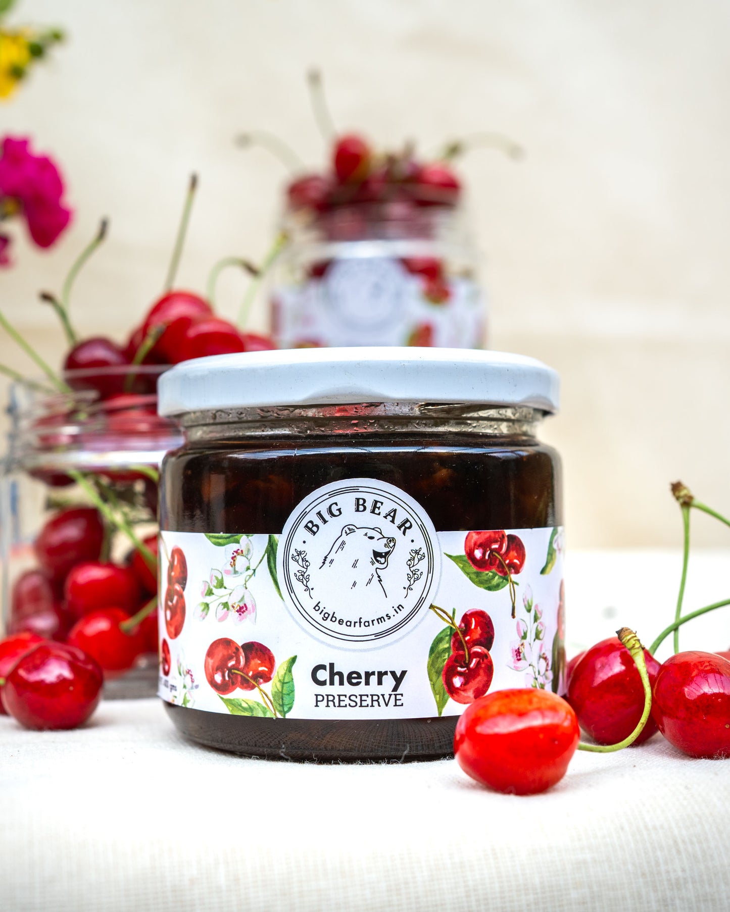 A jar of Big Bear Farms Cherry Preserve sits on a white fabric surface, surrounded by fresh red cherries. The background features a soft-focus second jar filled with cherries and blurred flowers.