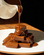 Caramel candies stacked on a speckled white plate, being drizzled with thick caramel sauce from a white pitcher against a dark background.