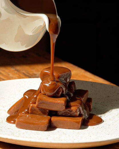 Caramel candies stacked on a speckled white plate, being drizzled with thick caramel sauce from a white pitcher against a dark background.