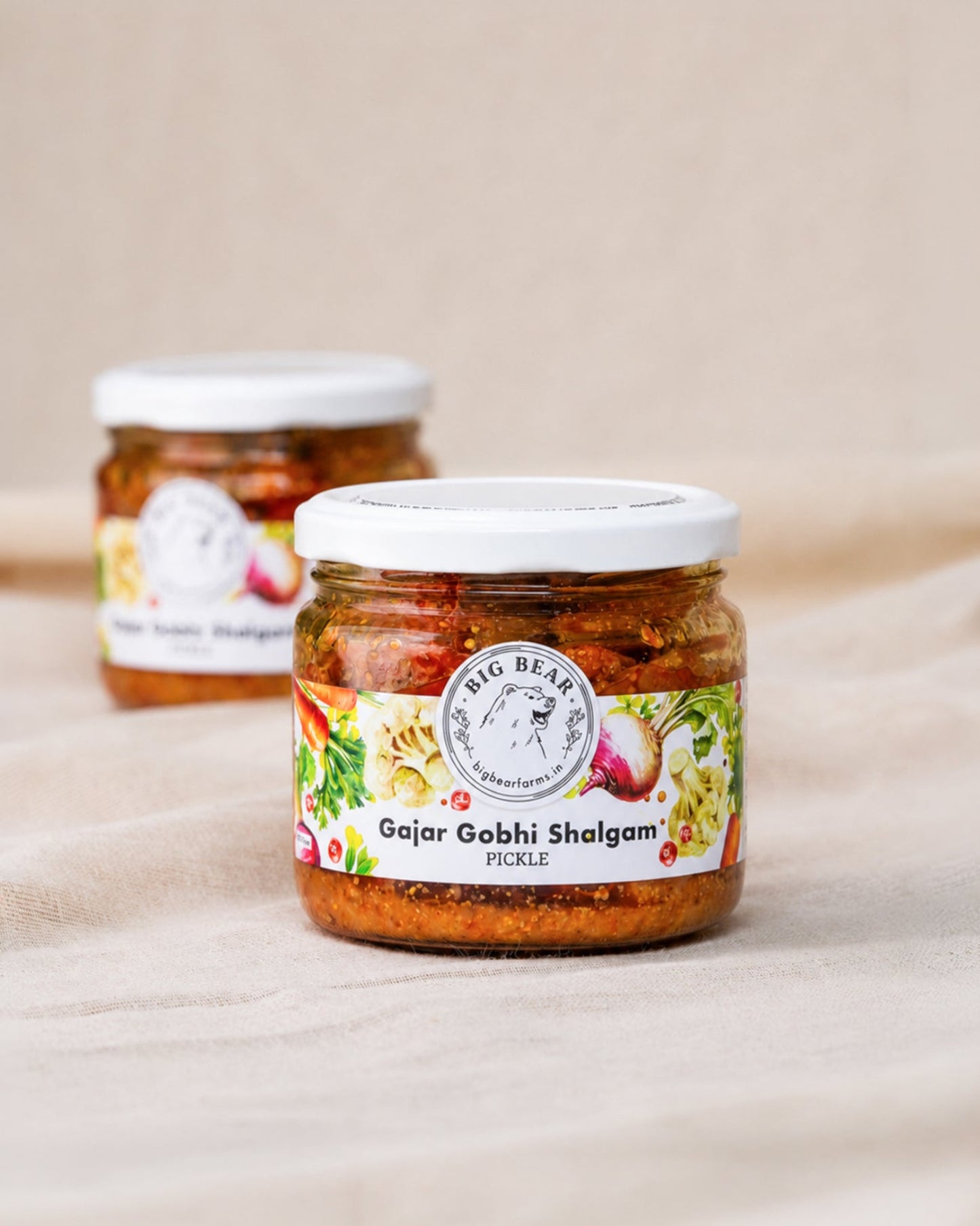 Two glass jars of Big Bear Pickles Gajar Gobhi Shalgam Pickle arranged on a beige fabric surface, with the front jar in sharp focus and the rear jar softly blurred. Carrot Radish Cauliflower pickle.