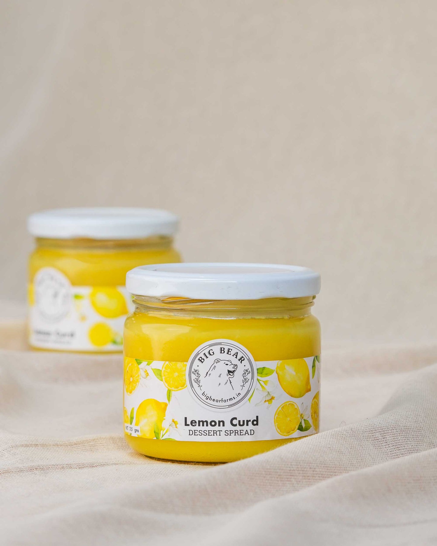 Two glass jars of Big Bear Farms
 Lemon Curd dessert spread, with the front jar in sharp focus on a draped beige fabric background.