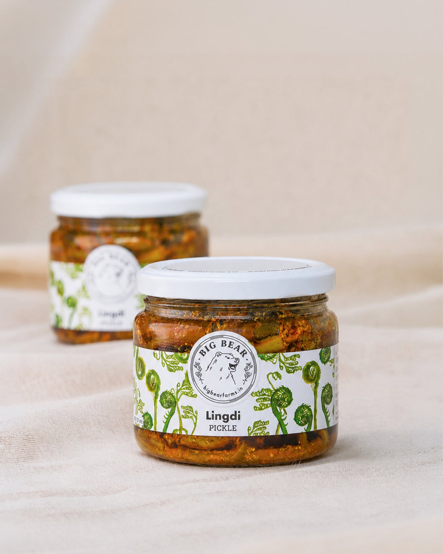 Two glass jars of Big Bear Farms Lingdi Pickle (fiddlehead fern pickle) on a neutral beige fabric background, with one jar in sharp focus in the foreground and the other softly blurred in the background.