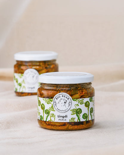 Two glass jars of Big Bear Farms Lingdi Pickle (fiddlehead fern pickle) on a neutral beige fabric background, with one jar in sharp focus in the foreground and the other softly blurred in the background.