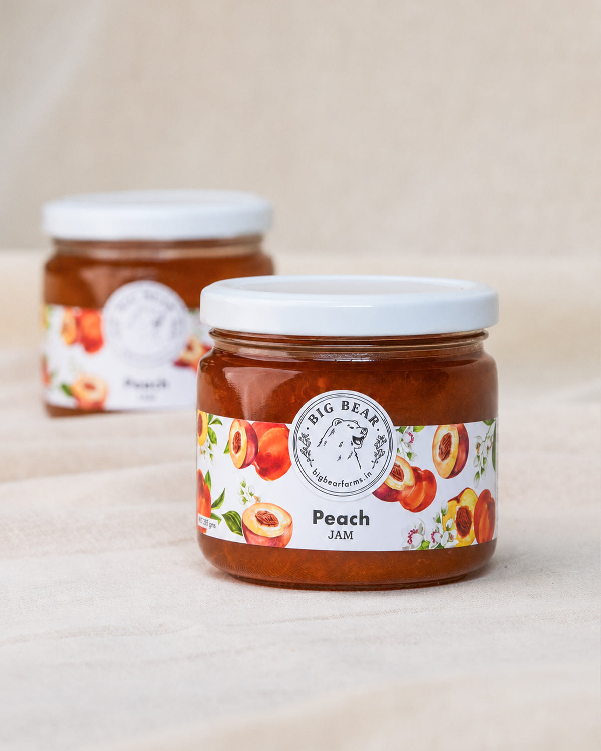 Two glass jars of Big Bear Farms Peach Jam on a light beige fabric surface, with the front jar in sharp focus and the rear jar softly blurred.