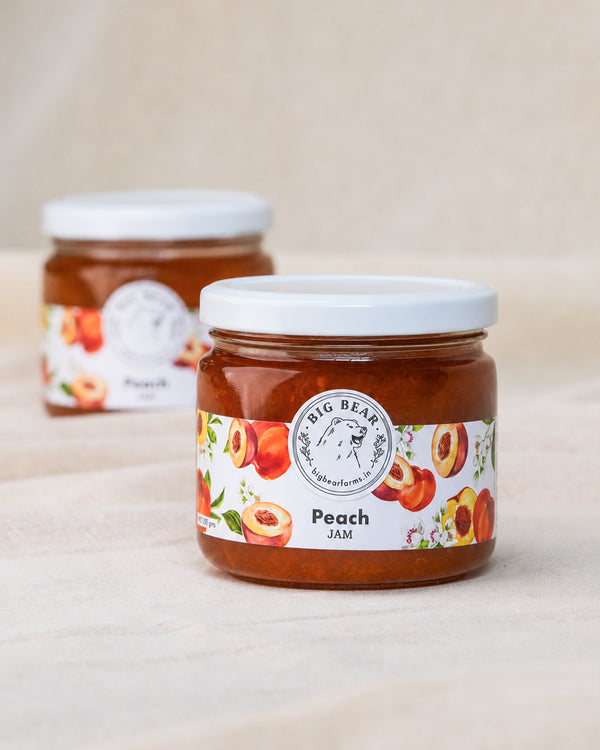 Two glass jars of Big Bear Farms Peach Jam on a light beige fabric surface, with the front jar in sharp focus and the rear jar softly blurred.