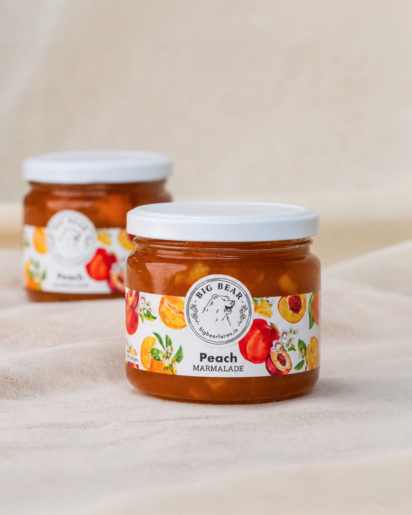Two glass jars of Big Bear Farms Peach Marmalade on a beige fabric background, with the front jar in sharp focus and the rear jar softly blurred.