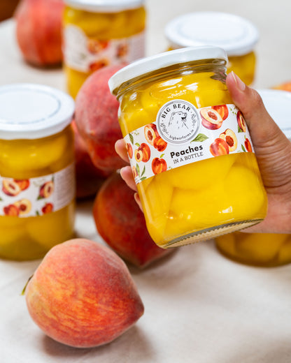 A hand holds a glass jar of Big Bear Farms Peaches in a bottle, surrounded by fresh peaches and other jars on a light neutral surface.