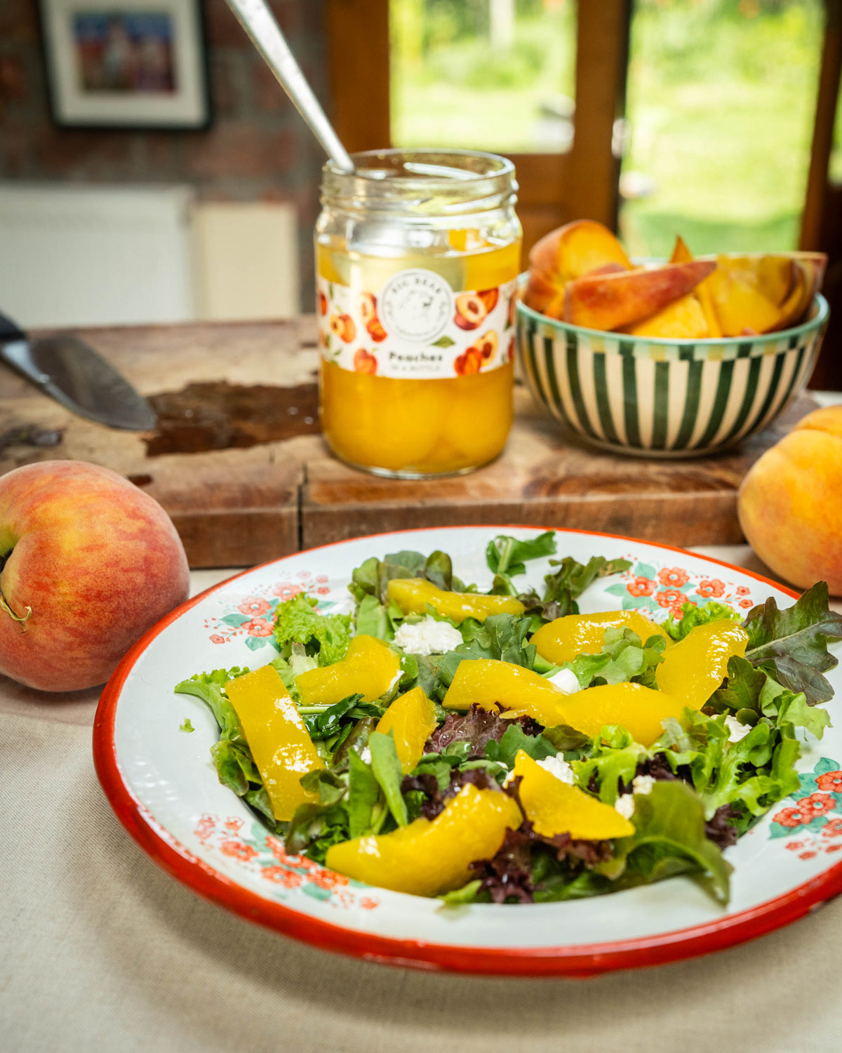 A jar of Big Bear Farms peach slices sits on a rustic wooden cutting board next to a bowl of peach slices, with a fresh peach salad in a white enamel plate with a red rim in the foreground.