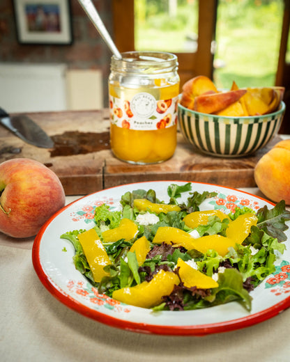 A jar of Big Bear Farms peach slices sits on a rustic wooden cutting board next to a bowl of peach slices, with a fresh peach salad in a white enamel plate with a red rim in the foreground.
