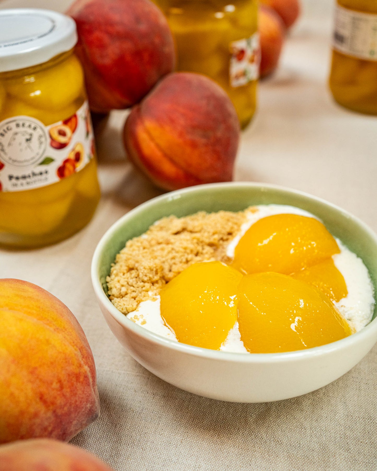 A bowl of yogurt topped with Big Bear Farms fresh peach slices and crumbled granola, with jars of Big Bear Farms peaches in a bottle and whole peaches arranged around it on a beige fabric surface.