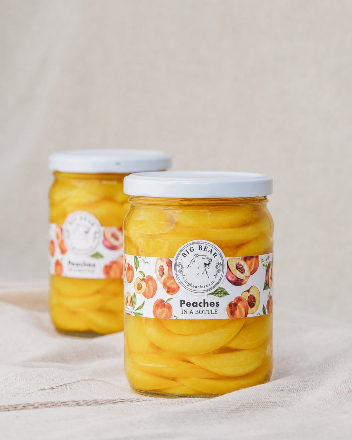 Two glass jars of Big Bear Peaches in a Bottle, filled with bright yellow peach slices and topped with white lids, set on a beige fabric surface.