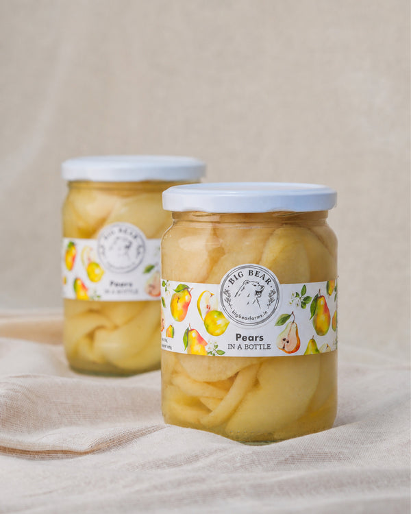 Two glass jars of Big Bear Farm Fresh Pears in a Bottle spread on a beige fabric surface. The front jar is in sharp focus, showing pale yellow pears submerged in liquid, with a white label featuring pears and a white lid.