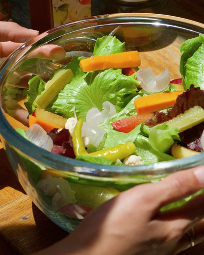 A bowl filled with a fresh salad and big bear farms fresh pickled vegetables, held by hands over a wooden table. The salad contains big bear farms fresh pickled vegetables orange carrot sticks, cucumber, green chillies, cauliflower, green lettuce and white onions.