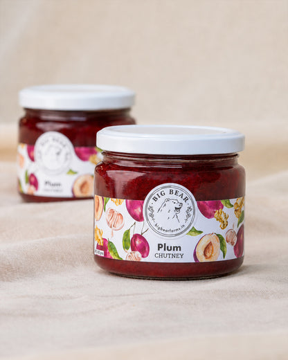 Two glass jars of Big Bear Farms Plum Chutney on a beige fabric background. The front jar is in sharp focus, showing the dark red chutney inside and a white label with fruit illustrations, while the second jar is softly blurred in the background.