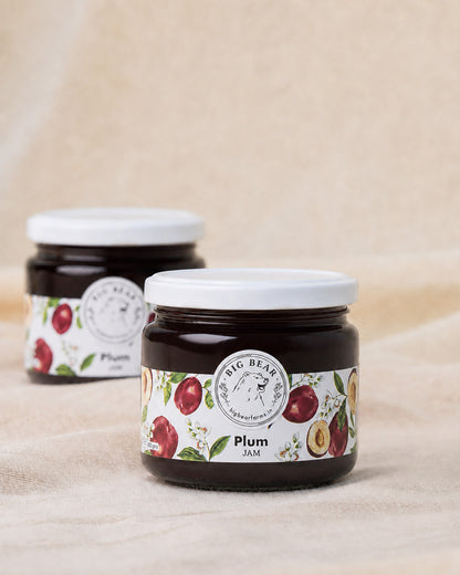 Two glass jars of Big Bear Farms Plum Jam on a beige fabric background, with one jar in sharp focus in the foreground and the other softly blurred in the background.
