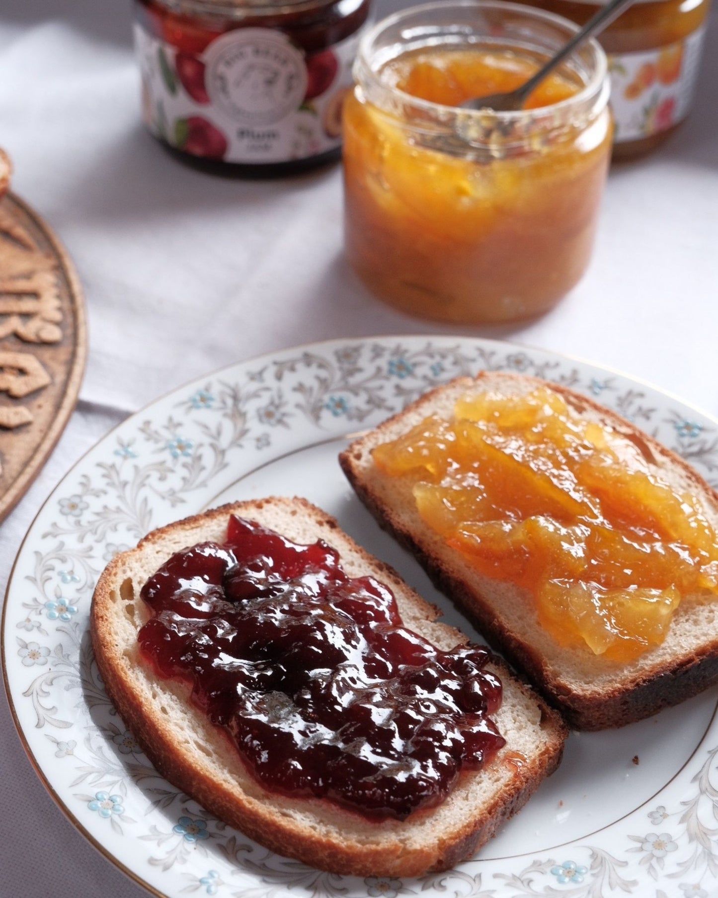 Two slices of toast on a white floral plate, one spread with Big Bear Farms plum jam and the other with Big Bear Farms marmalade.