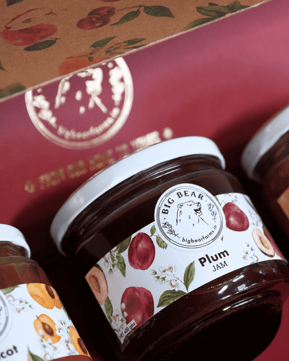 A close-up of a gift box containing jars of Big Bear Farm Plum Jam, with one jar fully visible and others partially cropped. The packaging features a deep red box with gold lettering and a kraft paper lid decorated with fruit illustrations.