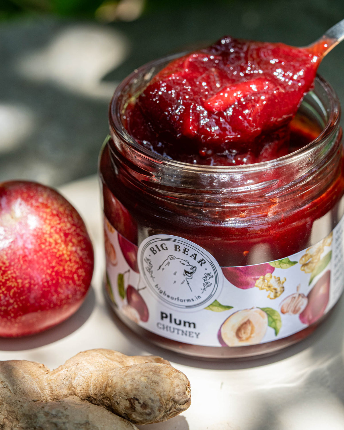 An open glass jar of Big Bear Farms Plum Chutney with a spoonful of thick red chutney showing the texture and pieces of ginger, styled with a whole plum and a piece of ginger root on a sunlit surface.