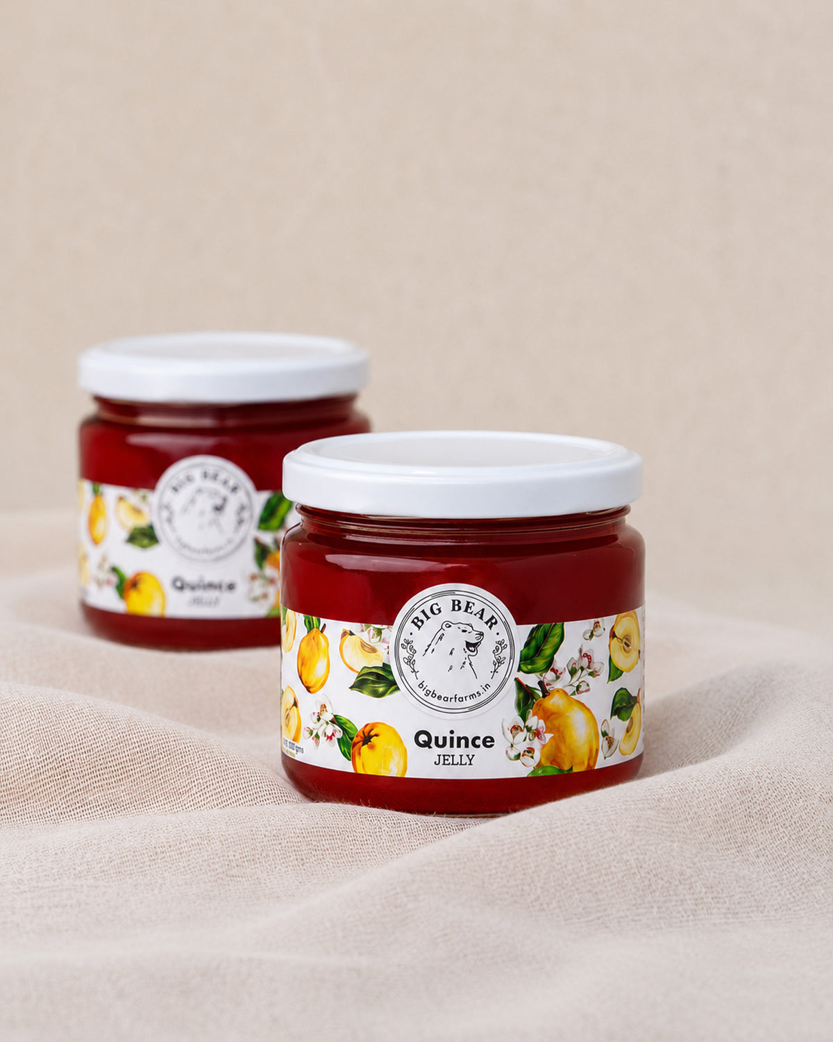 Two glass jars of Big Bear Farms Quince Jelly with white lids, displayed on a beige fabric surface with a matching background.