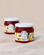 Two glass jars of Big Bear Farms Quince Jelly with white lids, displayed on a beige fabric surface with a matching background.