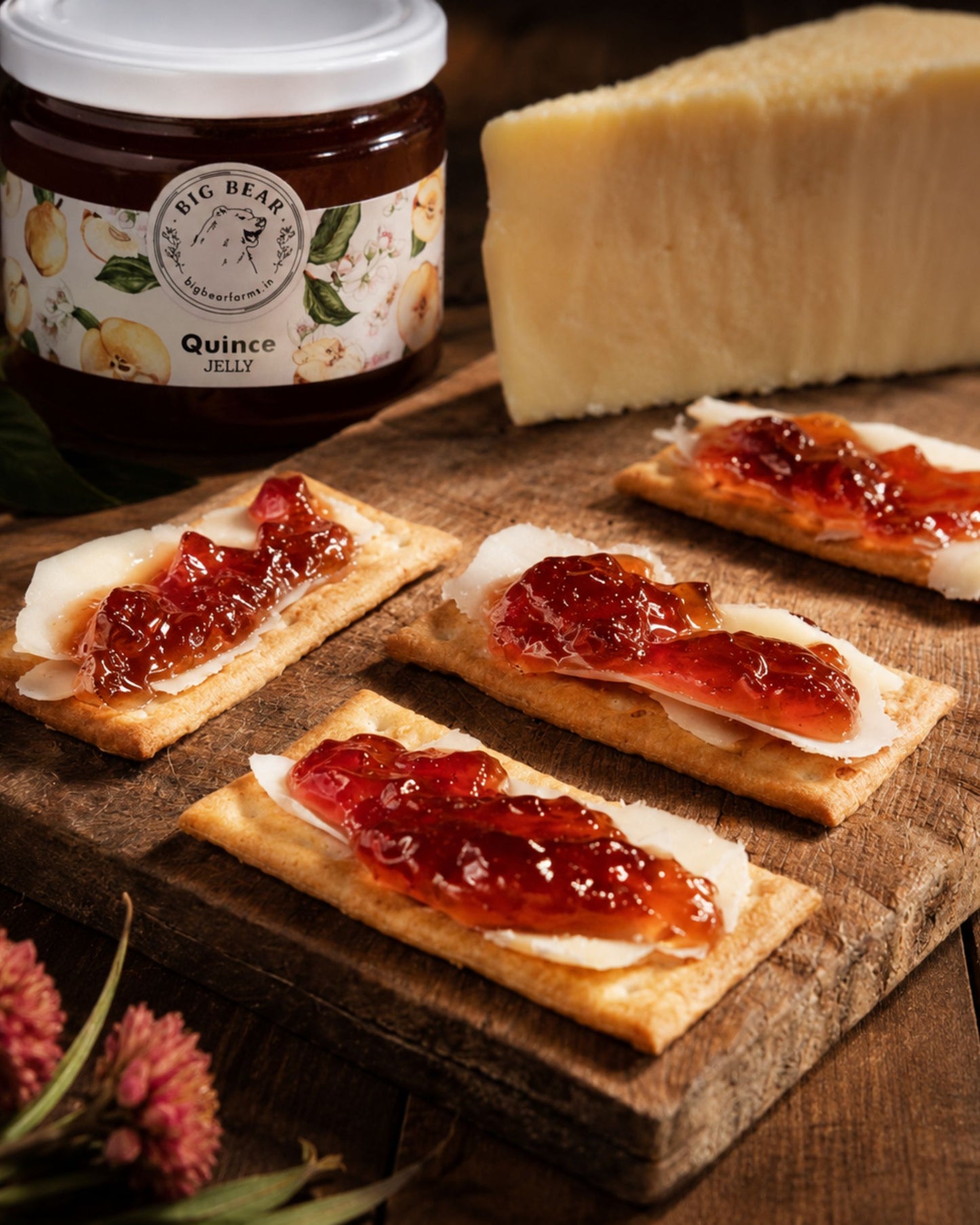 A jar of Big Bear Farms Quince Jelly is shown with several open-faced crackers topped with Big Bear Farms Quince Jelly on slices of cheese on a wooden board, alongside a wedge of cheese.