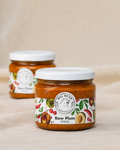 Two glass jars of Big Bear Farms Raw Plum Pickle with white lids, displayed on a beige fabric background.