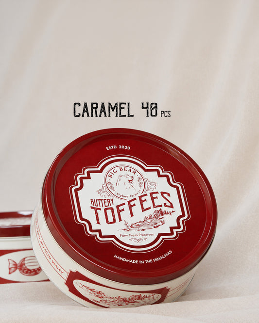 A round tin of 40 Big Bear Farms Buttery Toffees in the Caramel flavor, shown on a draped cream fabric background with a second tin partially visible behind it. The label is deep red and white with the brand name 'Big Bear' and product name 'Buttery Toffees' clearly visible.