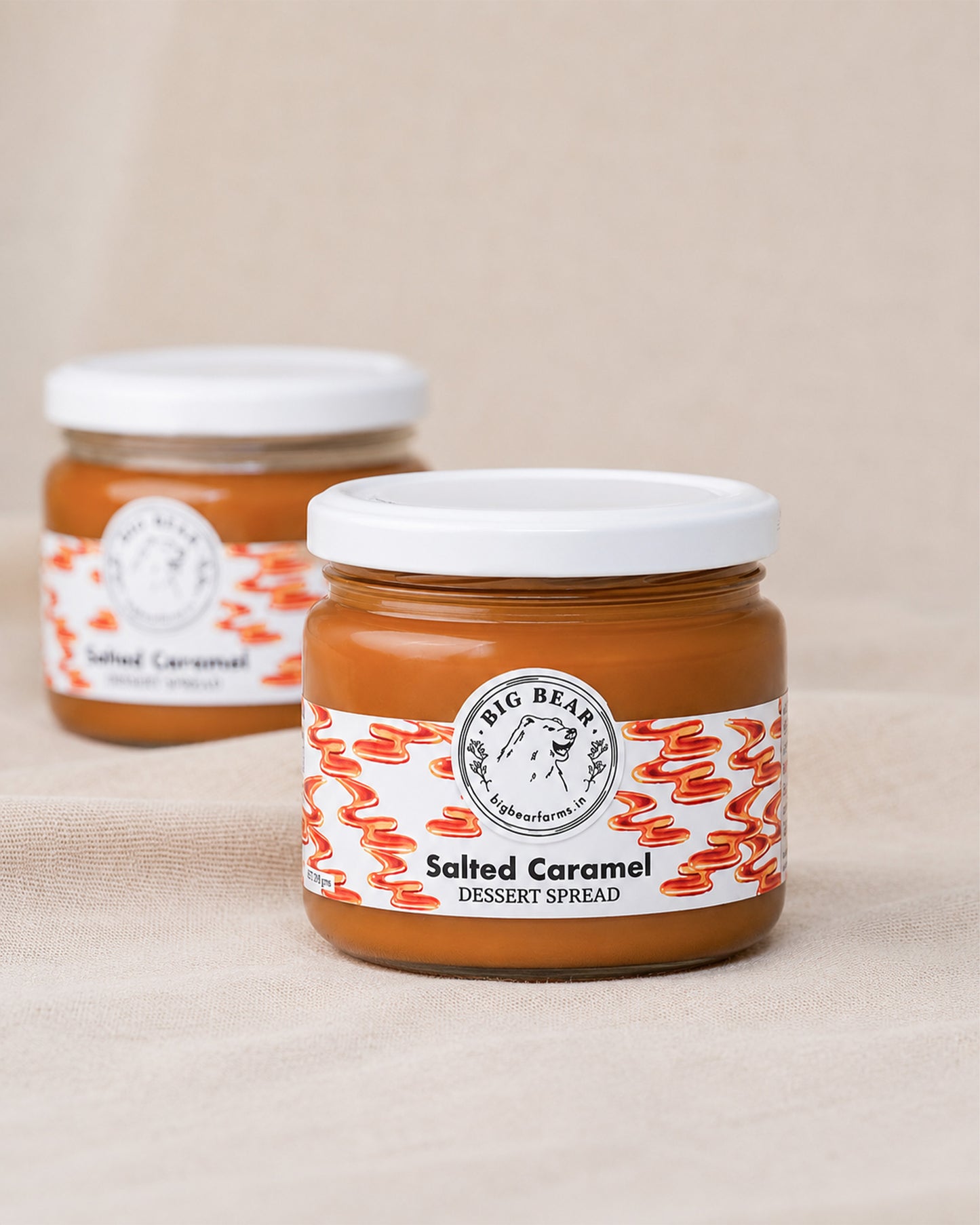 Two glass jars of Big Bear Farms Salted Caramel Dessert Spread, with the front jar in sharp focus and the second jar softly blurred in the background, set against a neutral beige fabric backdrop.