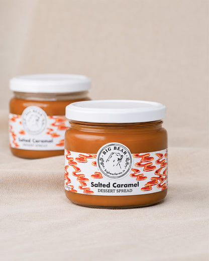 Two glass jars of Big Bear Farms Salted Caramel Dessert Spread, with the front jar in sharp focus and the second jar softly blurred in the background, set against a neutral beige fabric backdrop.